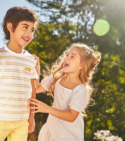 Par de irmãos, um menino e uma menina, dão risada. Eles estão ao ar livre, num jardim ensolarado. Ambos vestem roupas infantis de cor clara da marca Carinhoso.