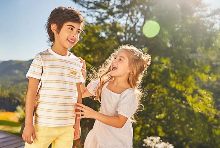 Par de irmãos, um menino e uma menina, dão risada. Eles estão ao ar livre, num jardim ensolarado. Ambos vestem roupas infantis de cor clara da marca Carinhoso.