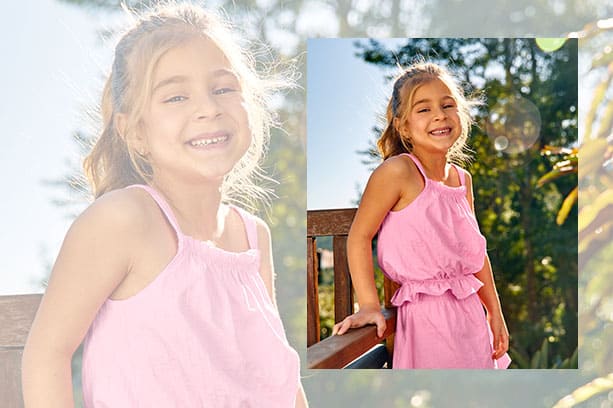 Menina loira ri para a câmera em foto ao ar livre. Ela veste conjunto infantil cor de rosa de regata cropped e shorts saia da Malwee Kids.