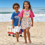 Crianças na praia usando roupas de verão. Menino negro de óculos usando blusa azul e short branco infantil. Menina branca usando jardineira infantil.