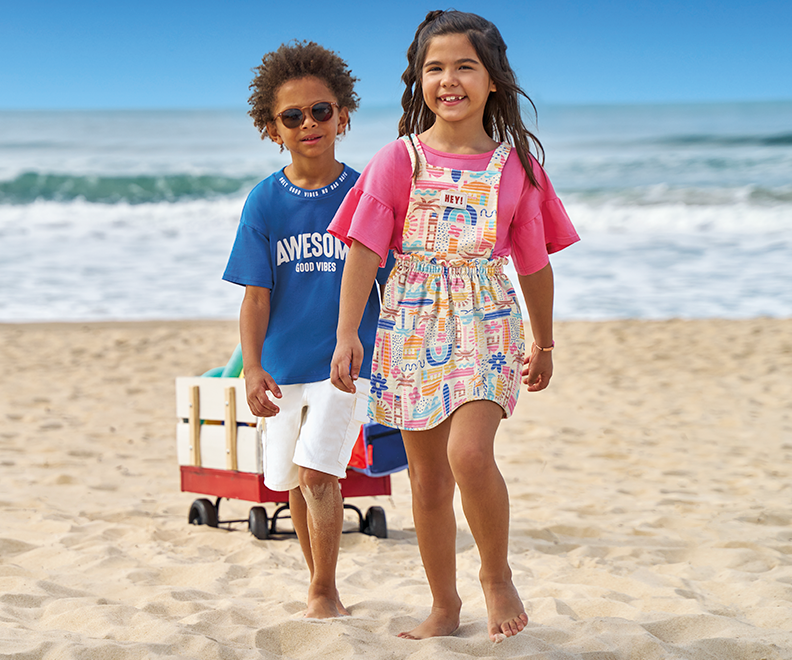 Crianças na praia usando roupas de verão. Menino negro de óculos usando blusa azul e short branco infantil. Menina branca usando jardineira infantil.