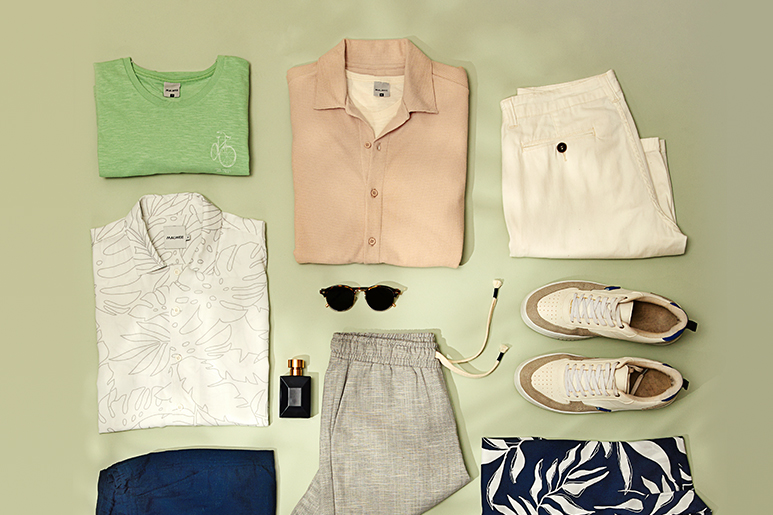 Diversas roupas e acessórios masculinas. Camisa verde, branca estampada e bege dobrada. Óculos, perfume e tênis. Bermuda branca e bermuda cinza dobrada em fundo verde. 