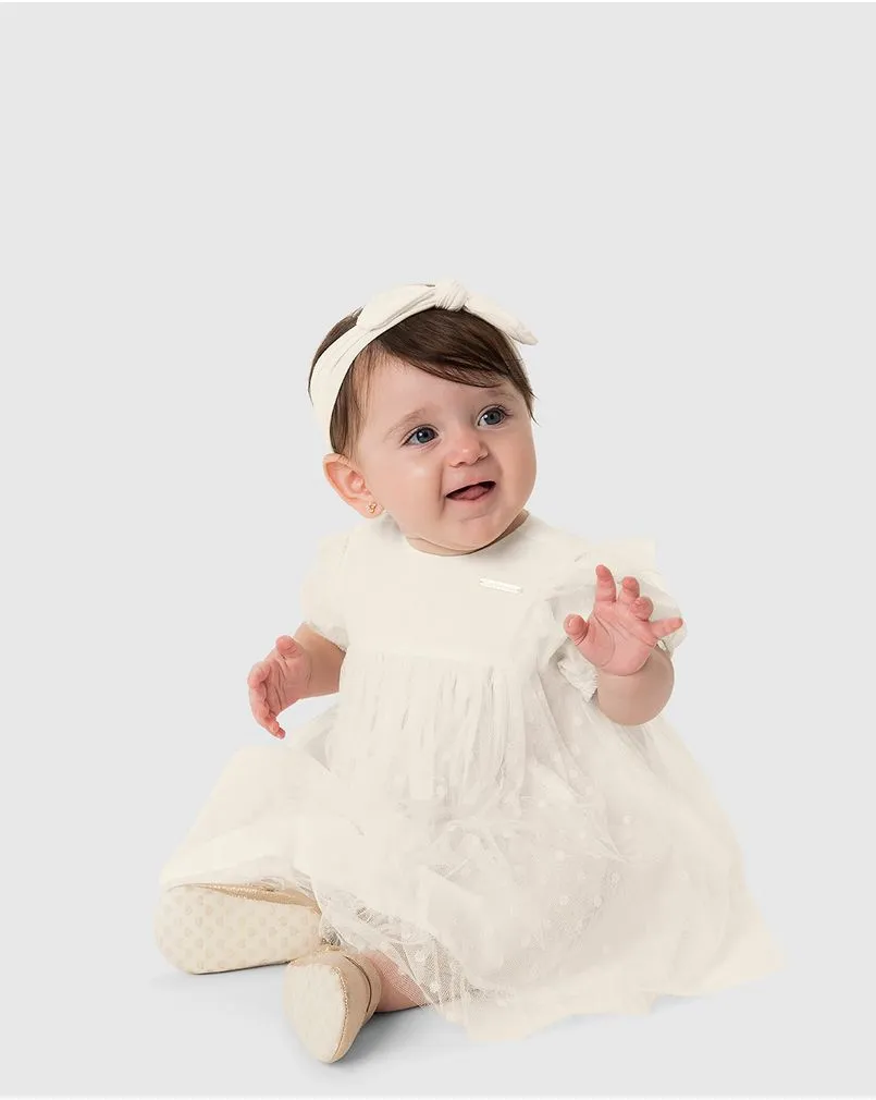 Bebê menina veste um conjunto de vestido e calcinha brancos para matizado da marca Carinhoso.