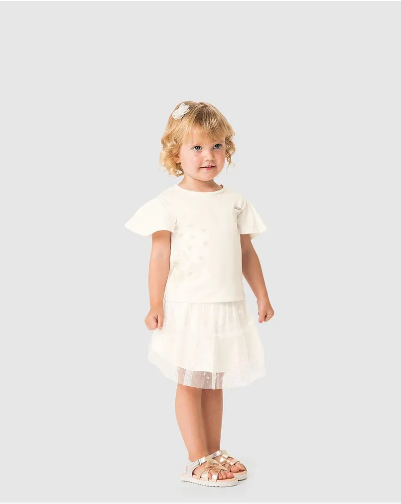Criança menina veste um conjunto branco de camiseta e saia em tule, para batizado, da marca Carinhoso.