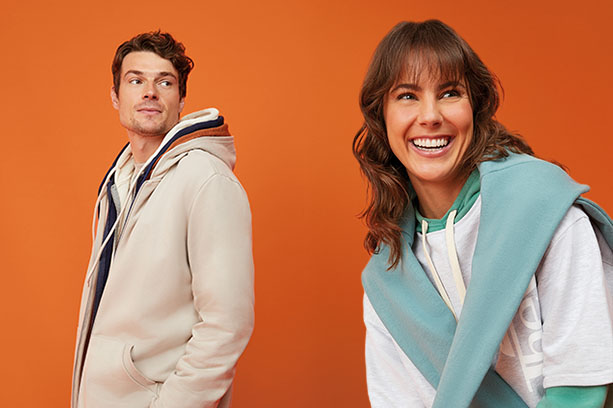 Modelo feminina sorridente veste um moletom feminino verde de capuz por baixo de uma camiseta básica cinza. Atrás dela, um modelo masculino olha para o lado, vestindo vários moletons masculinos da Malwee, em sobreposição.