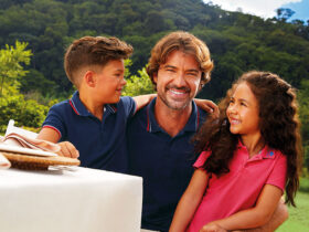 Uma família brasileira posa para uma foto durante almoço ao ar livre. O pai e seu casal de filhos vestem peças da coleção Dia dos Pais 2025 da Malwee. O filho criança veste uma camisa polo infantil azul marinho; o pai veste uma camisa polo masculina azul marinho, enquanto a filha veste um vestido polo infantil cor de rosa.