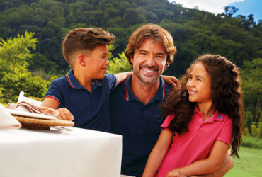 Uma família brasileira posa para uma foto durante almoço ao ar livre. O pai e seu casal de filhos vestem peças da coleção Dia dos Pais 2025 da Malwee. O filho criança veste uma camisa polo infantil azul marinho; o pai veste uma camisa polo masculina azul marinho, enquanto a filha veste um vestido polo infantil cor de rosa.