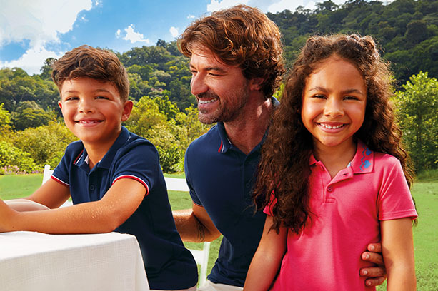 Uma família brasileira posa para uma foto durante um almoço ao ar livre. O filho criança veste uma camisa polo infantil da Malwee Kids. O pai, um homem adulto branco de cabelos castanhos, veste uma camisa polo masculina azul marinho da Malwee. A filha criança veste um vestido com gola polo cor de rosa da Malwee Kids.