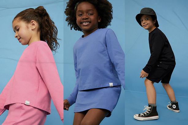 Crianças, em frente a um fundo azul, vestem roupas de moletom nas cores rosa, azul caribe e preto. As peças têm etiqueta da Malwee Kids.