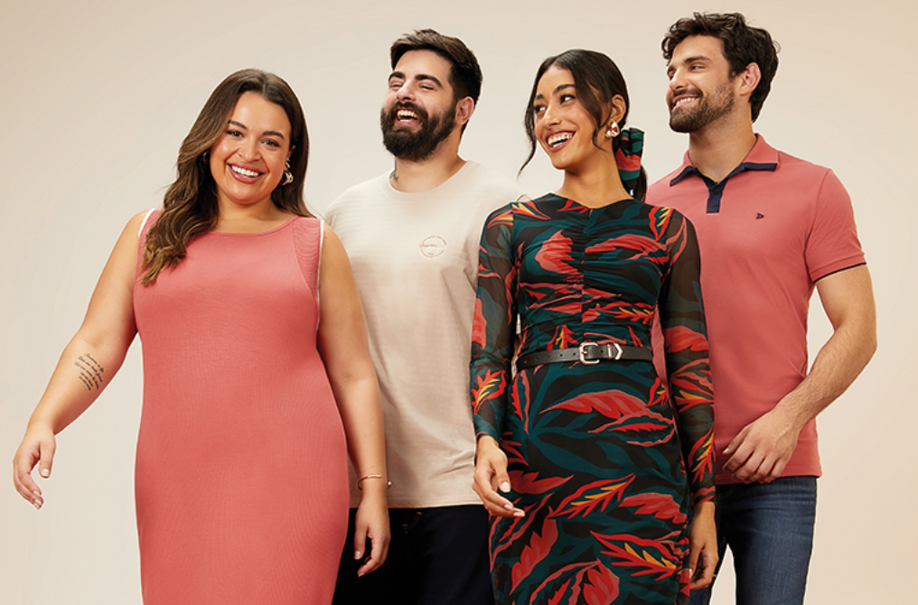 Quatro pessoas sorrindo em um estúdio com fundo bege. À esquerda, uma mulher de 32 anos com cabelos castanhos ondulados e pele clara veste um vestido rosa sem mangas. Ao lado dela, um homem de 35 anos com barba e cabelos castanhos veste uma camiseta bege. Ao lado dele, uma mulher de 29 anos com cabelos pretos presos em um rabo de cavalo e pele morena veste um vestido de manga comprida com estampa floral em tons de verde, vermelho e preto, e um cinto preto fino. À direita, um homem de 33 anos com barba e cabelos castanhos veste uma camisa polo rosa com detalhes em azul marinho e jeans azul. Todos estão sorrindo e olhando para frente. Em estilo de campanha publicitária.