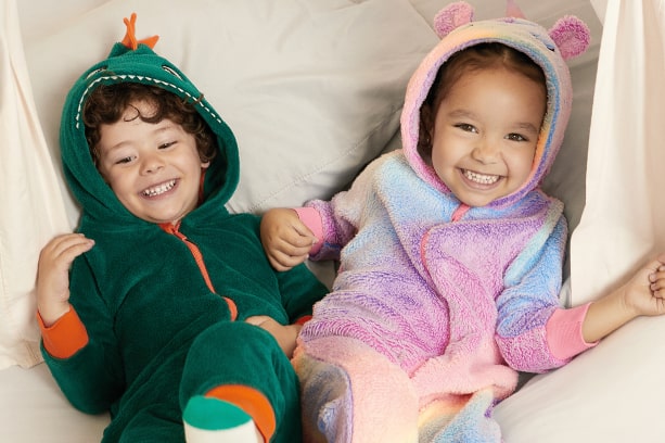 Duas crianças sorrindo em uma cama com lençóis brancos. À esquerda, um menino latino de 5 anos com cabelos castanhos cacheados veste um macacão de fleece verde com capuz de dinossauro. À direita, uma menina de 6 anos com cabelos pretos lisos veste um macacão de fleece colorido com capuz de unicórnio.