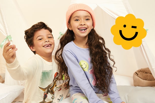 Duas crianças sorrindo em um ambiente interno com um fundo bege claro. À esquerda, um menino latino de 7 anos com cabelos castanhos cacheados veste um pijama amarelo claro com estampa de dinossauro. À direita, uma menina asiática de 8 anos com cabelos castanhos longos veste um pijama lilás com estampa de carinha sorridente. A menina usa uma touca rosa. No canto superior direito, um adesivo amarelo com uma carinha sorridente piscando