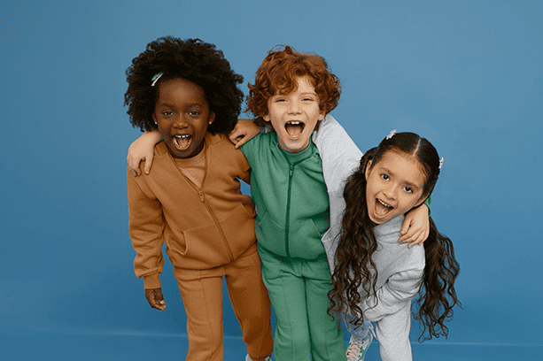 Três modelos crianças fazem careta e posam abraçados, olhando para a câmera. Eles vestem conjuntos de moletons básicos da Malwee Kids, coloridos, quentinhos e confortáveis.