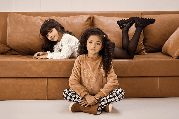 Duas meninas posam em um sofá de couro. Uma delas está deitada no sofá, de bruços, olhando para a câmera. A segunda modelo está sentada no chão, olhando para a câmera. Ambas vestem looks de moda infantil premium da Carinhoso.