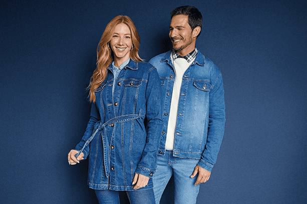 Look all jeans: Um casal de modelos, um homem e uma mulher, posam olhando para a câmera em frente a um fundo azul. Os dois vestem roupas em jeans azul da Malwee.