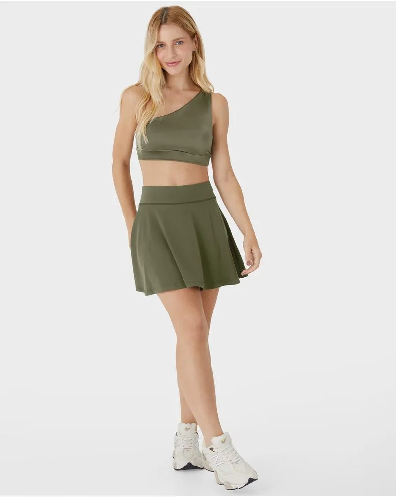 Modelo loira da marca Enfim Active veste um conjunto de academia verde militar, com top de um ombro só e saia plissada.