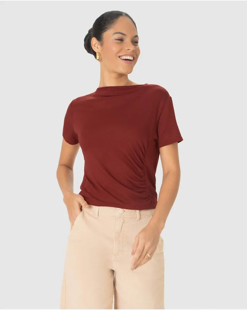 Modelo morena da marca Malwee, com cabelo em coque, veste uma camiseta de manga curta vermelha com decote canoa e tecido franzido na região lateral esquerda do tronco. 