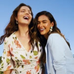 Modelos da Malwee posam sorridentes em um parque ensolarado. Uma delas está vestindo um vestido branco florido, e a outra veste um blazer azul céu.