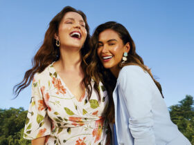 Modelos da Malwee posam sorridentes em um parque ensolarado. Uma delas está vestindo um vestido branco florido, e a outra veste um blazer azul céu.