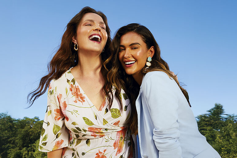 Modelos da Malwee posam sorridentes em um parque ensolarado. Uma delas está vestindo um vestido branco florido, e a outra veste um blazer azul céu.