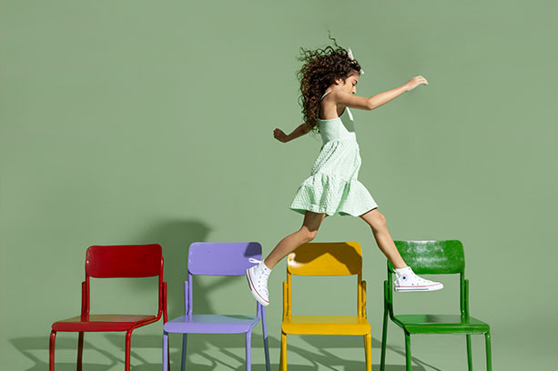 Menina com vestido de festa verde Carinhoso brinca em cadeiras coloridas 
