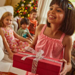 Criança de cabelo castanho e vestido malwee kids se prepara para presentear a amiga, uma criança loira também vestida com vestido florido da malwee kids, em comemoração de Natal.