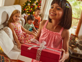 Criança de cabelo castanho e vestido malwee kids se prepara para presentear a amiga, uma criança loira também vestida com vestido florido da malwee kids, em comemoração de Natal.