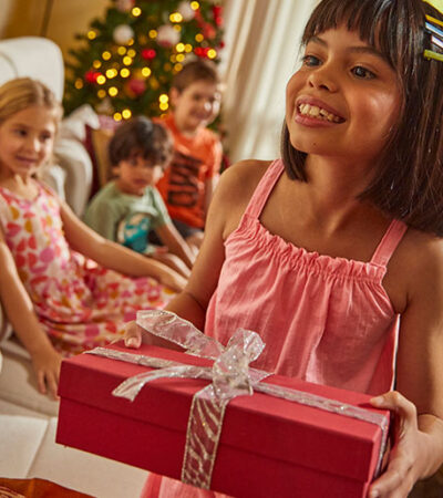 Criança de cabelo castanho e vestido malwee kids se prepara para presentear a amiga, uma criança loira também vestida com vestido florido da malwee kids, em comemoração de Natal.
