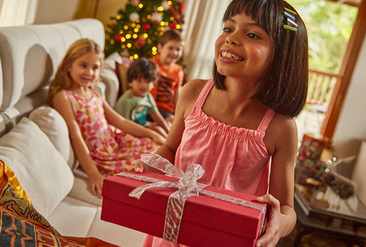 Criança de cabelo castanho e vestido malwee kids se prepara para presentear a amiga, uma criança loira também vestida com vestido florido da malwee kids, em comemoração de Natal.