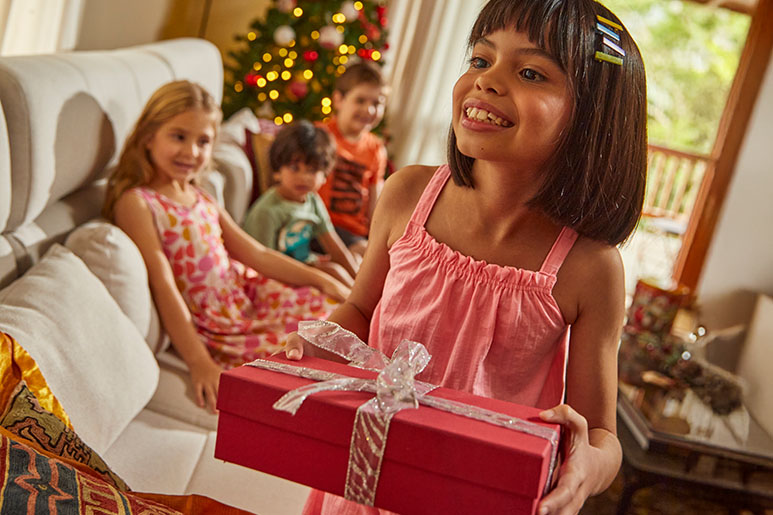 Criança de cabelo castanho e vestido malwee kids se prepara para presentear a amiga, uma criança loira também vestida com vestido florido da malwee kids, em comemoração de Natal.