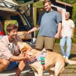 Família de três homens, todos vestindo Malwee, se prepara para viagem junto ao seu cachorro.